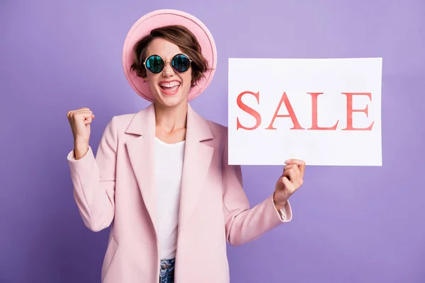 Porträt von nett überrascht fröhliche junge Frau hält in der Hand Verkauf Banner tragen Mütze Brille isoliert auf pastelllila Hintergrund — Stockfoto