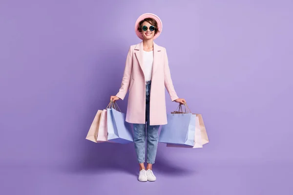 Фотографія повного розміру весела позитивна молода жінка тримає покупки одягають окуляри рожеве пальто джинси взуття ізольовані на фіолетовому фоні — стокове фото