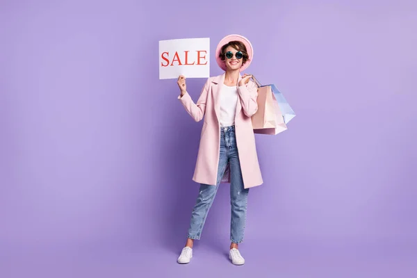 Фото позитивної красивої дівчини, що тримає покупки продажу плакат одягати окуляри пальто джинси взуття ізольовано на фіолетовому фоні — стокове фото