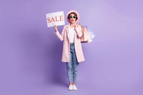 Foto in voller Größe von überrascht nettes Mädchen halten Käufe Verkauf Plakat tragen Brille Mantel Jeans Schuhe isoliert auf lila Hintergrund — Stockfoto