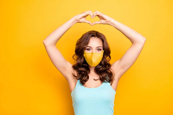 Retrato de namorada morena fazendo amor símbolo figura do coração com os dedos sobre a cabeça usando máscara de tecido amarelado isolado no fundo amarelo — Fotografia de Stock