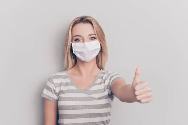 Close-up portret van vrouw tonen duim tot het dragen van wit gezicht masker geïsoleerd op grijs gekleurde achtergrond — Stockfoto