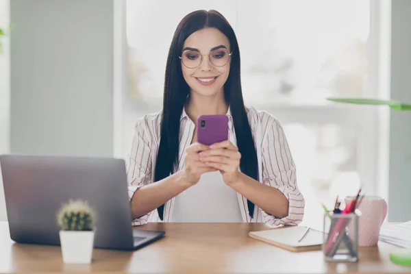 Фото умной улыбающейся деловой женщины, сидящей за столом и смотрящей на руки гаджета, болтающей смс на рабочем месте — стоковое фото