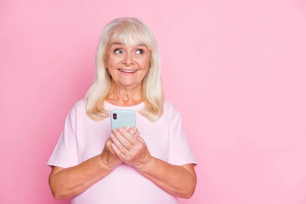 Meraklı büyükannenin resmi akıllı telefonu elinde tutuyor boş alana bakıyor pastel pembe arka planda mutlu bir şekilde izole edilmiş gülümsüyor — Stok fotoğraf