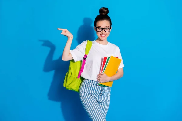 Classroom number one here. Positive college student girl hold books rucksack point finger copyspace demonstrate courses ads promo wear white denim isolated bright shine color background