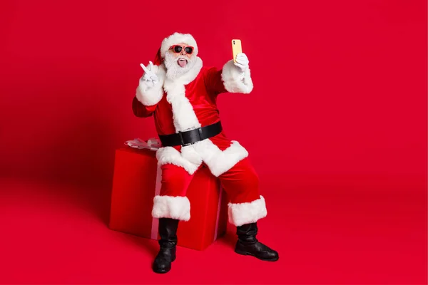 Emekli yaşlı bir adamın fotoğrafı beyaz sakallı, hediye kutusu V-tabelası gösteriyor ki özçekim portresi öne çıkıyor, dil dışarı çıkıyor, x-mas Noel Baba ceketi, eldiven kemer, güneş gözlüğü kapağı, izole edilmiş kırmızı arka plan. — Stok fotoğraf