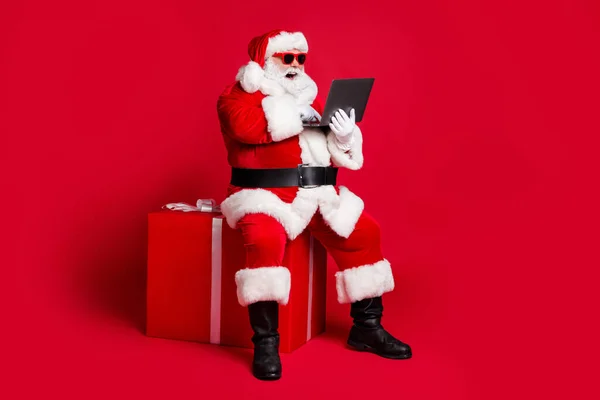 Volledige lengte foto van gepensioneerde oude man witte baard zitten giftbox houden laptop bestellingen online dragen x-mas santa kostuum handschoen riem zonneglas kap boot geïsoleerde rode kleur achtergrond — Stockfoto