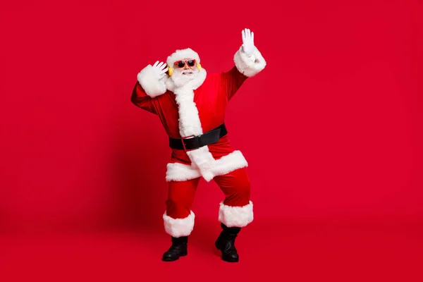 Pleine longueur photo de retraité vieil homme barbe grise dansant insouciant porter santa costume gants manteau ceinture lunettes de soleil casque sans fil écouteurs bottes en cuir noir isolé fond de couleur rouge — Photo