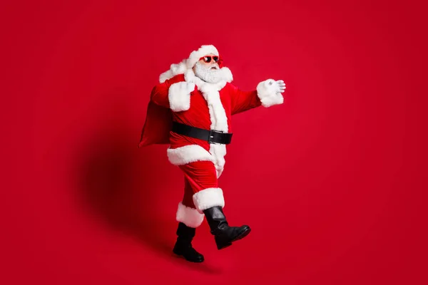 Foto de perfil de longitud completa de hombre viejo retirado barba gris paseo oso bolsa regalo responsable abuelo prisa antes de la medianoche desgaste traje de Santa botas de gafas de sol aislado color rojo fondo —  Fotos de Stock