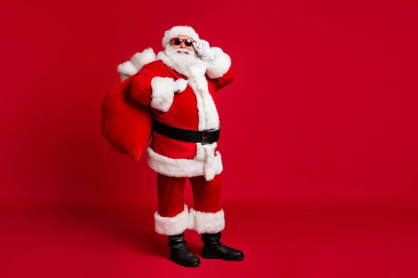 Emekli yaşlı bir adamın tam boy fotoğrafı gri sakallı ayı gözlüklü çanta iyi çocuklar Noel Baba kostümlü eldiven ceket, güneş gözlüklü başlık siyah botlar izole edilmiş kırmızı arka plan. — Stok fotoğraf