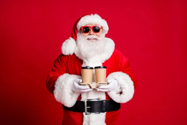 Fotografie důchodce starý muž šedé vousy držet kávu jít barista dodat vám dva šálky pohodlné ranní nošení x-mas Santa kostým rukavice kabát pás sluneční sklo čepice izolované červené barvy pozadí — Stock fotografie