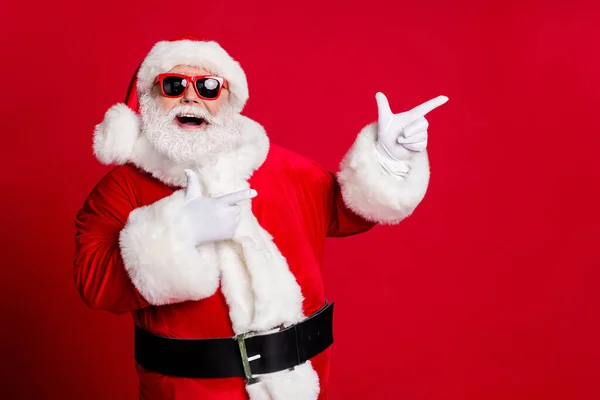 Foto di pensionato vecchio uomo grigio barba bocca aperta sorriso dita dirette spazio vuoto dimostrare migliore opzione indossare Santa x-mas guanti costume cappotto occhiali da sole copricapi isolato colore rosso sfondo — Foto Stock