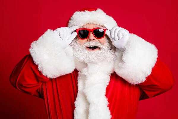 Cant believe my eyes. Closeup portrait photo of retired old man grey beard open mouth smiling hold glasses see north pole wear santa costume sunglass headwear isolated red color background