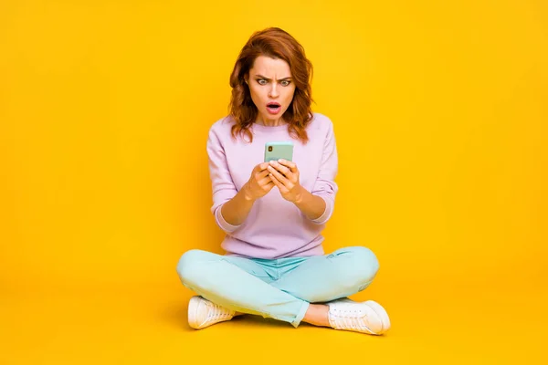 フルボディ写真ショックを受けた女性が座って足を交差使用スマートフォンを読む恐ろしいソーシャルメディアニュース感銘を受けた悲鳴を着用ピンクの靴ジャンパー隔離された黄色の色の背景 — ストック写真