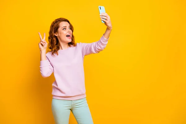 Hello Fénykép vicces csinos hölgy tartsa telefon kezét, hogy szelfi mutatja v-jel szimbólum mondván hi kopás rózsaszín pulóver zöld nadrág elszigetelt sárga háttér — Stock Fotó