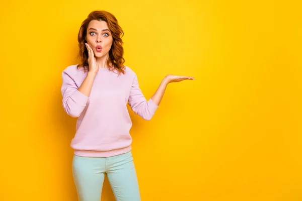 Foto van grappige krullende dame verhogen nieuwigheid product op open palm gek breken nieuws zwarte vrijdag prijzen dragen casual roze trui groene broek geïsoleerde gele kleur achtergrond — Stockfoto