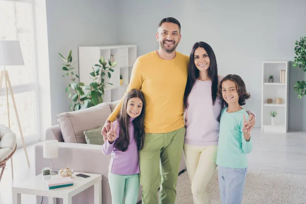 Foto van het hele gezin vier leden verzamelen van twee kleine kinderen knuffel show v-teken glimlach dragen kleurrijke trui broek in de woonkamer binnen — Stockfoto