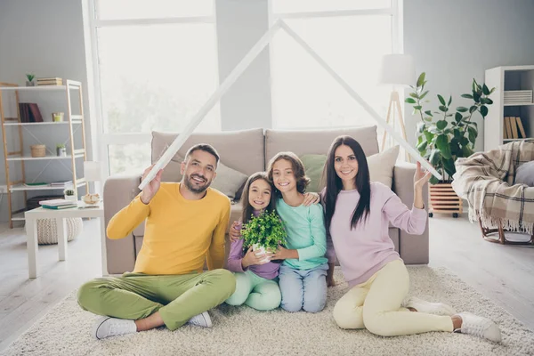 Foto da família completa quatro membros sentar-se chão pais segurar papel telhado crianças flowerpot usar calças de camisola coloridas na sala de estar dentro de casa — Fotografia de Stock