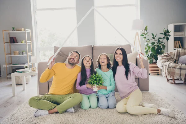 Фотографія повної сім'ї чотирьох членів сидять батьки на підлозі тримають дах над головою дітей квітка носить барвисті светр штани у вітальні в приміщенні — стокове фото