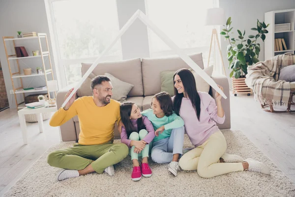 全家人四个人的照片两个小孩妈妈爸爸抱着纸屋顶坐在地毯上穿着五颜六色的套头衫在客厅里 — 图库照片