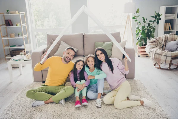 Фотографія повної сім'ї чотирьох членів сідла мама тато тримає паперовий дах сидить на підлозі посмішка камера носити барвистий джемпер у вітальні в приміщенні — стокове фото