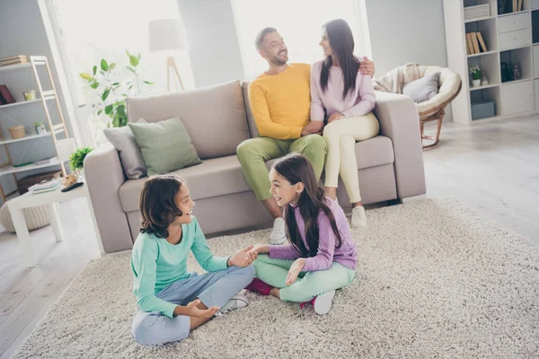 Фотографія повної сім'ї чотирьох членів сидять на дивані двоє маленьких дітей мають чат одягнені барвисті штани-перемички у вітальні в приміщенні — стокове фото