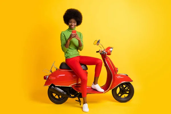 붉은 스쿠터에 앉아 있는 젊은 흑인 여성의 사진, 선명 한 노란색 콜드 배경에 고립 된 녹색 점퍼를 입고 메시지를 보내는 핸드폰을 들고 앉아 있는 미국 여성의 사진 — 스톡 사진