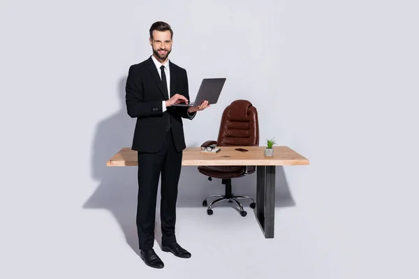 Полное фото красивого делового парня держать ноутбук руки болтать коллеги хорошее настроение стоять современный офис носить черный пиджак рубашка брюки костюм изолированный серый фон — стоковое фото