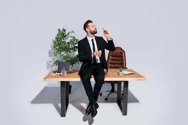 Ganzes Foto von gutaussehenden Geschäftsmann sitzen Tisch gute Laune halten Seifenblasen Flasche weht tragen Blazer Hemd Krawatte Hose Schuhe Kostümanzug isoliert grauen Hintergrund — Stockfoto