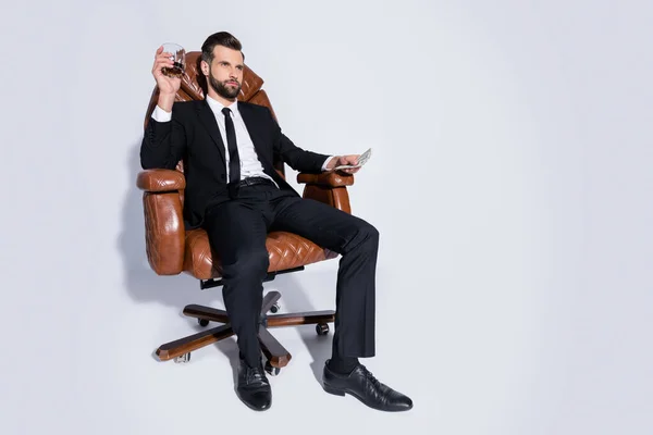 Ganzes Foto von schönen Business-Kerl sitzen Stuhl halten Pack Geld Dollar trinken Luxus teuer Scotch bereit zahlen tragen schwarze Blazer Hose Krawatte Hemdschuhe Anzug isoliert grauen Hintergrund — Stockfoto