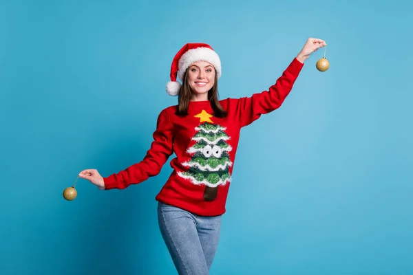 Портретне фото усміхненої задоволеної дівчини в смішному червоному різдвяному вбранні, що тримає демонстрацію декоративних золотих різдвяних кульок, ізольованих на синьому кольорі — стокове фото