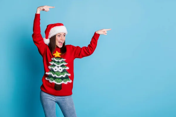 Photo of charming pretty lady raise hands direct look indicate empty space open mouth wear santa x-mas headwear red ornamented pullover jeans isolated blue color background — Stock Photo, Image