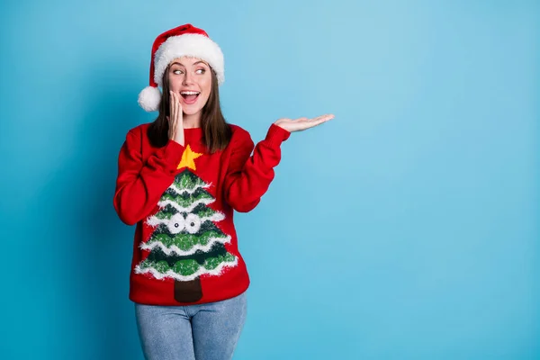 Photo of charming pretty lady hold look empty space amazed open mouth hand cheek wear santa x-mas headwear red ornamented sweater jeans isolated blue color background — Stock Photo, Image
