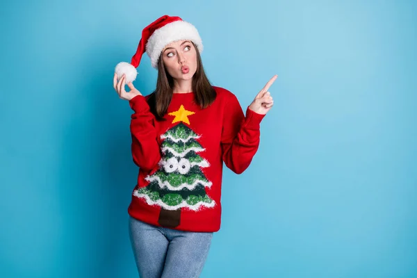 Foto de linda senhora bonita direta indicar dedo olhar para cima espaço vazio jogar pompom desgaste santa x-mas headwear vermelho ornamentado camisola jeans isolado azul cor fundo — Fotografia de Stock