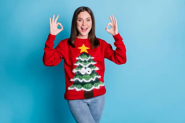 Photo of positive girl in christmas tree pullover sweater show jolly holly okay sign x-mas discounts promo wear denim jeans isolated over blue color background — Stock Photo, Image