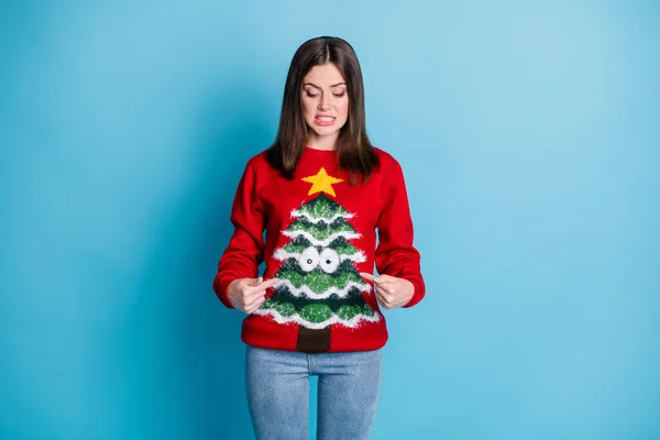 Worst gift idea ever. Photo portrait of young brunette disgusted with red sweater with tree star googly eyes pointing two fingers at self isolated on pastel light blue colored background