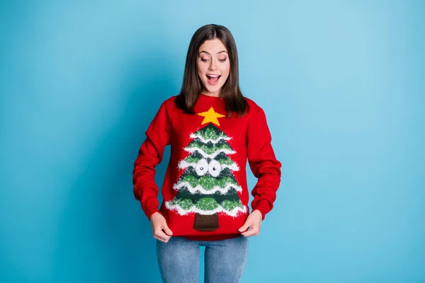 Photo portrait of surprised girl pulling ugly christmas sweater with hands smiling looking down isolated on pastel light blue colored background — Stock Photo, Image