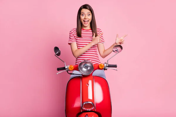 Foto portret van verrast meisje zitten op rode bromfiets wijzend naar lege ruimte glimlachend vrolijk geïsoleerd op pastel roze kleur achtergrond — Stockfoto