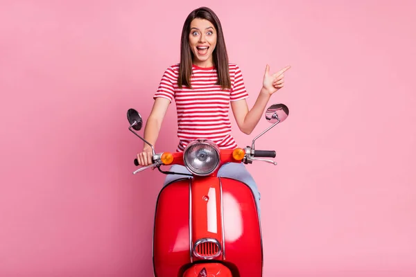 붉은 오토바이를 타고 빈 공간을 가리키며 파스텔 핑크 색 배경에 고립 된 입을 벌리고 웃고 있는 행복 한 소녀의 사진 — 스톡 사진