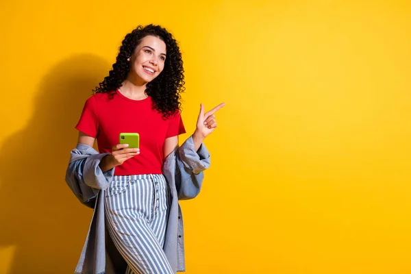 Bild av flicka hålla telefon direkt pekfinger ser tomt utrymme slitage randiga jeans röd t-shirt jacka isolerad gul färg bakgrund — Stockfoto