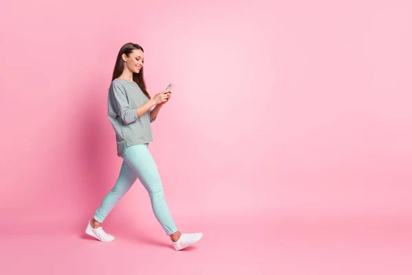 Full storlek profil foto av optimistiska söt ung kvinna kommer titta telefon bära blå jeans sneakers skjorta isolerad på rosa färg bakgrund — Stockfoto