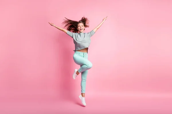 Foto de tamaño completo de optimista chica de pelo largo agradable saltar manos hasta usar camisa pantalones azules zapatillas blancas aisladas en el fondo de color rosa — Foto de Stock