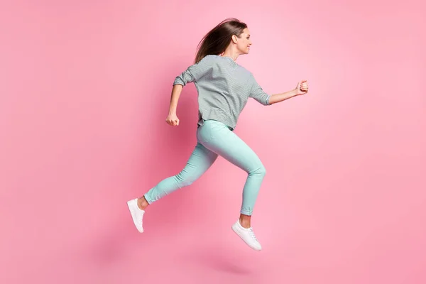 Teljes méretű profil fotó édes hosszú haj lány ugrás futás kéz ököl viselni ing kék nadrág fehér tornacipő elszigetelt rózsaszín háttér — Stock Fotó
