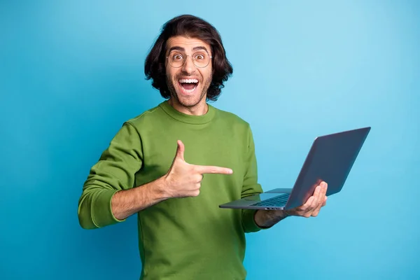 Foto van grappige opgewonden man houden netbook geven wijsvinger dragen brillen groene trui geïsoleerde blauwe kleur achtergrond — Stockfoto
