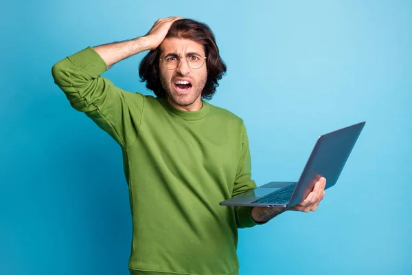 Foto van raadselachtige probleemjongen houden computer hand hoofd open mond slijtage specs groene trui geïsoleerde blauwe kleur achtergrond — Stockfoto
