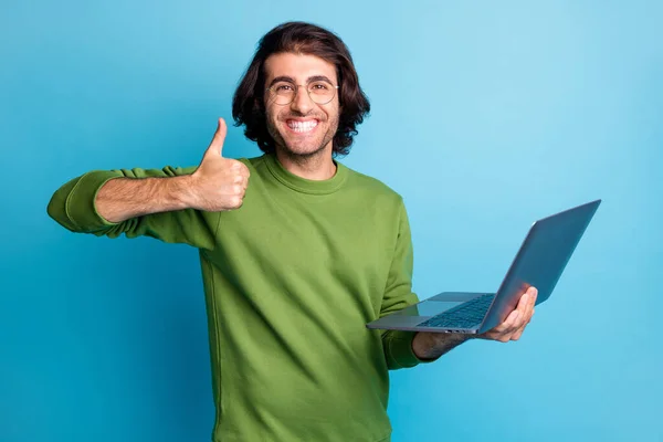 Фото молодого бородатого хлопця тримає комп'ютер, щоб підняти великий палець вгору, показує зелений пуловер ізольований синій колір фону — стокове фото