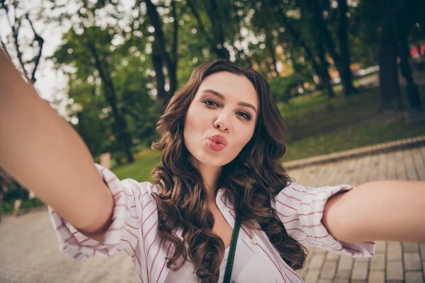 Foto retrato de una chica joven y bonita con el pelo largo y ondulado tomando selfie enviando un beso de aire con labios rellenos en el parque de la ciudad —  Fotos de Stock