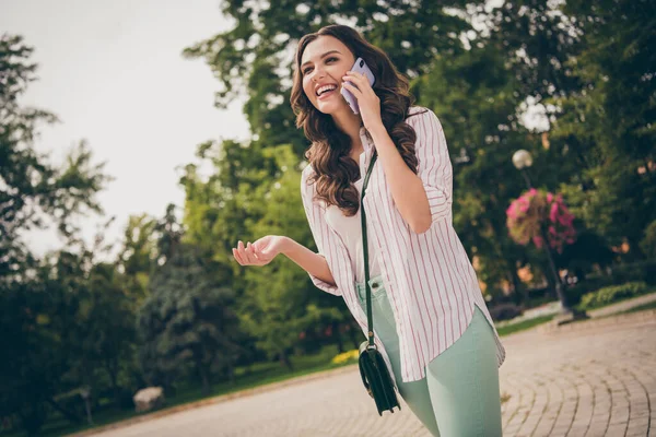 예쁜 여자가 핸드폰으로 웃으면서 평상복을 입고 녹색 도시 공원을 걷고 있는 사진 — 스톡 사진