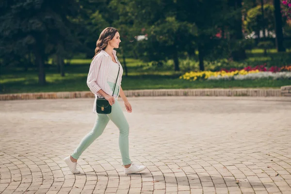 Full size profil fotografie roztomilé brunetky dáma chodit park aktivní život nosit pastel zelené džíny bílá vrchol pruhované košile taška přes rameno venku — Stock fotografie
