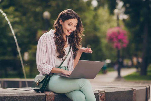 Foto da senhora morena otimista bonito sentar perto da fonte falando no laptop desgaste pastel jeans camisa bolsa de ombro superior no parque ao ar livre — Fotografia de Stock